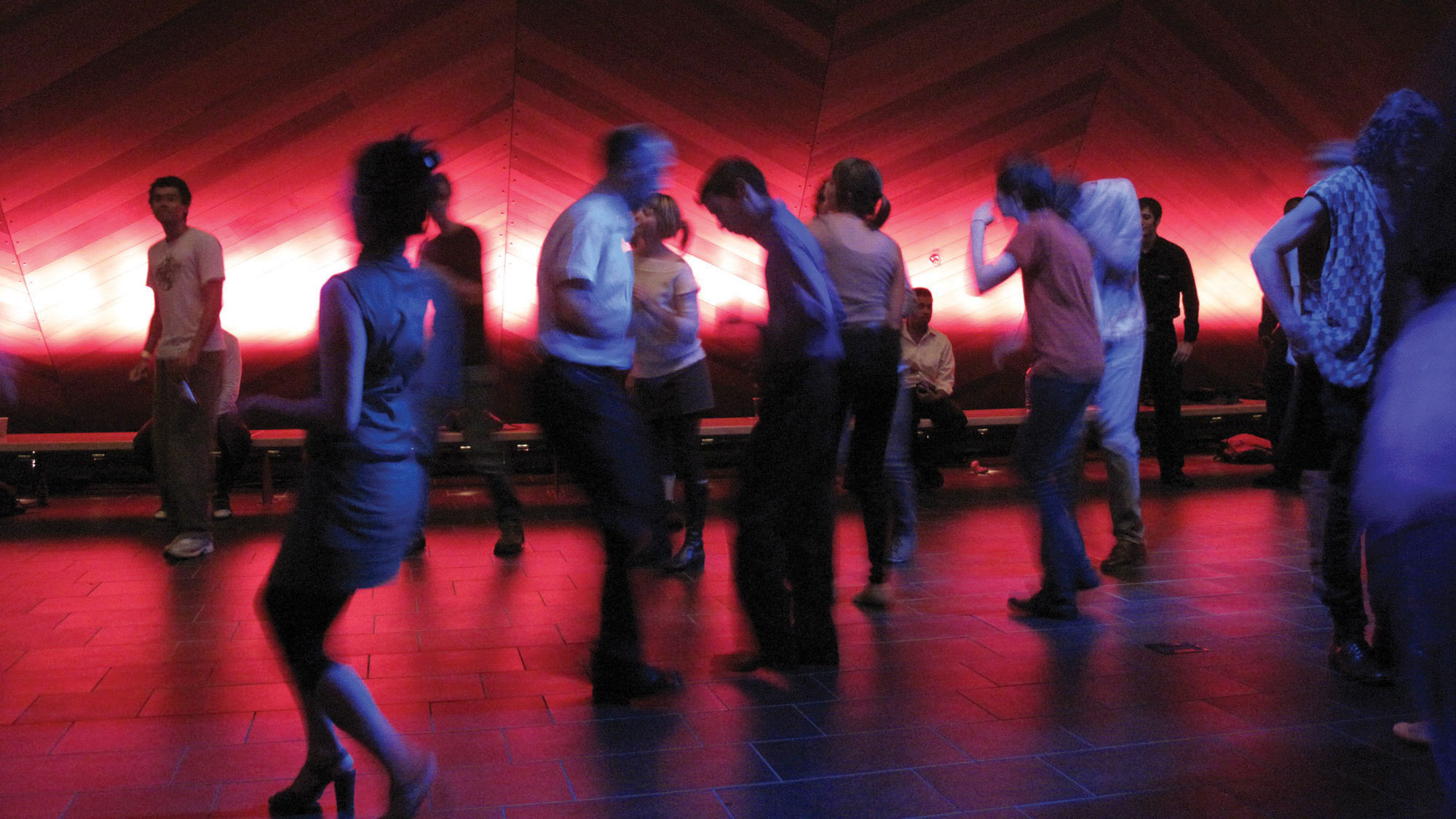 A group of about fifteen people blurred in dance and lit in red and blue light in front of the wooden hull of the EMPAC concert hall. 