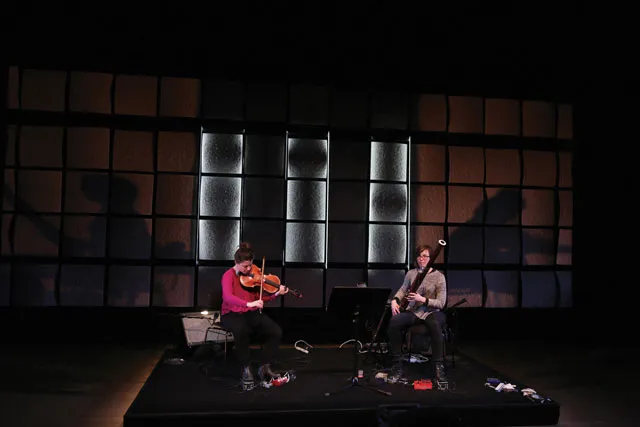 Two female musicians playing a viola and a bassoon on a small stage infant of a wall of tan acoustic tiles. 