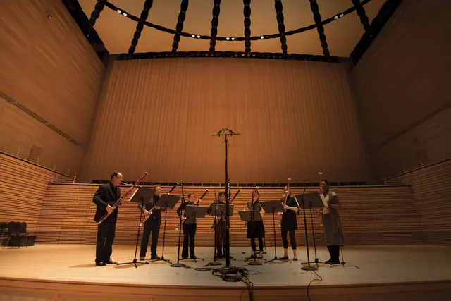 Seven bassoonists with music stands standing in a semi circle on the concert hall stage. 