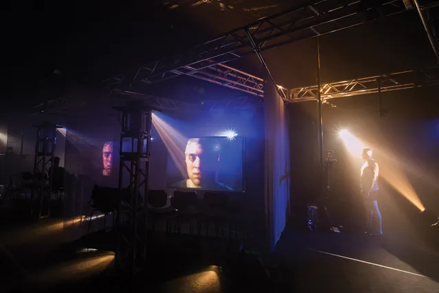 a male standing off to the right in a beam of theatrical light as his face is projecting on to two adjacent screens. 