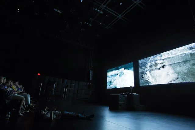 Two screens projecting scratched images of human eyes to a seated audience in a black theater. 