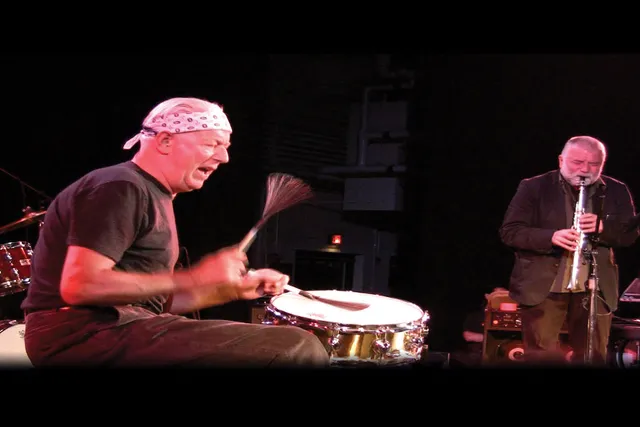 A white man wearing a bandana tied around his head playing the drums alongside another white man with a gray beard playing soprano sax. 
