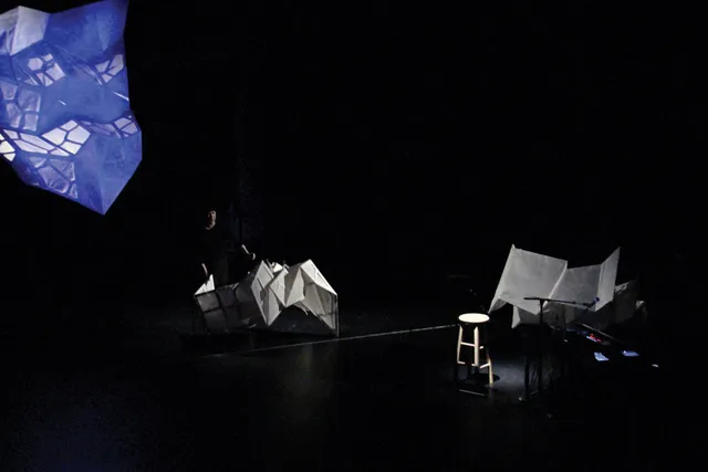 A woman on a dark stage amongst three large, white geometric sculptures. 