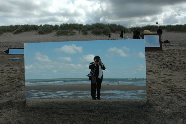 Agnes Varda taking a picture of herself in a large mirror placed on a beach. 