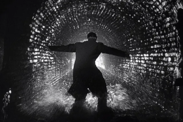 A man wearing a long coat with arms outstretched, touching each side of a cobblestone tunnel. 