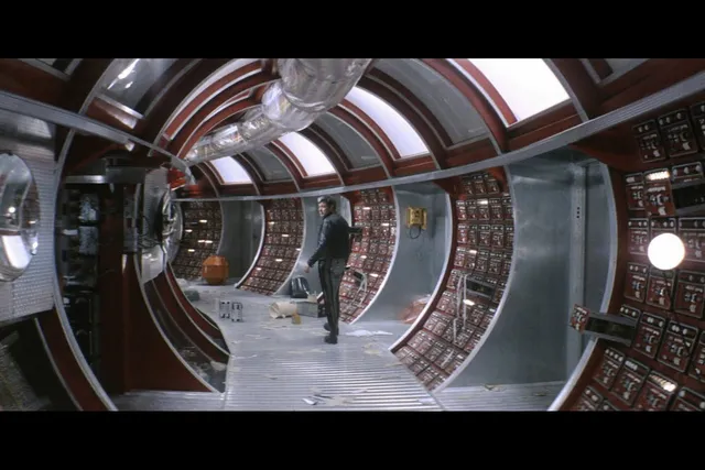 A man standing inside a messy hallway of a fictional spaceship. The steel gray and burgundy tunnel is lined in various nobs and buttons.