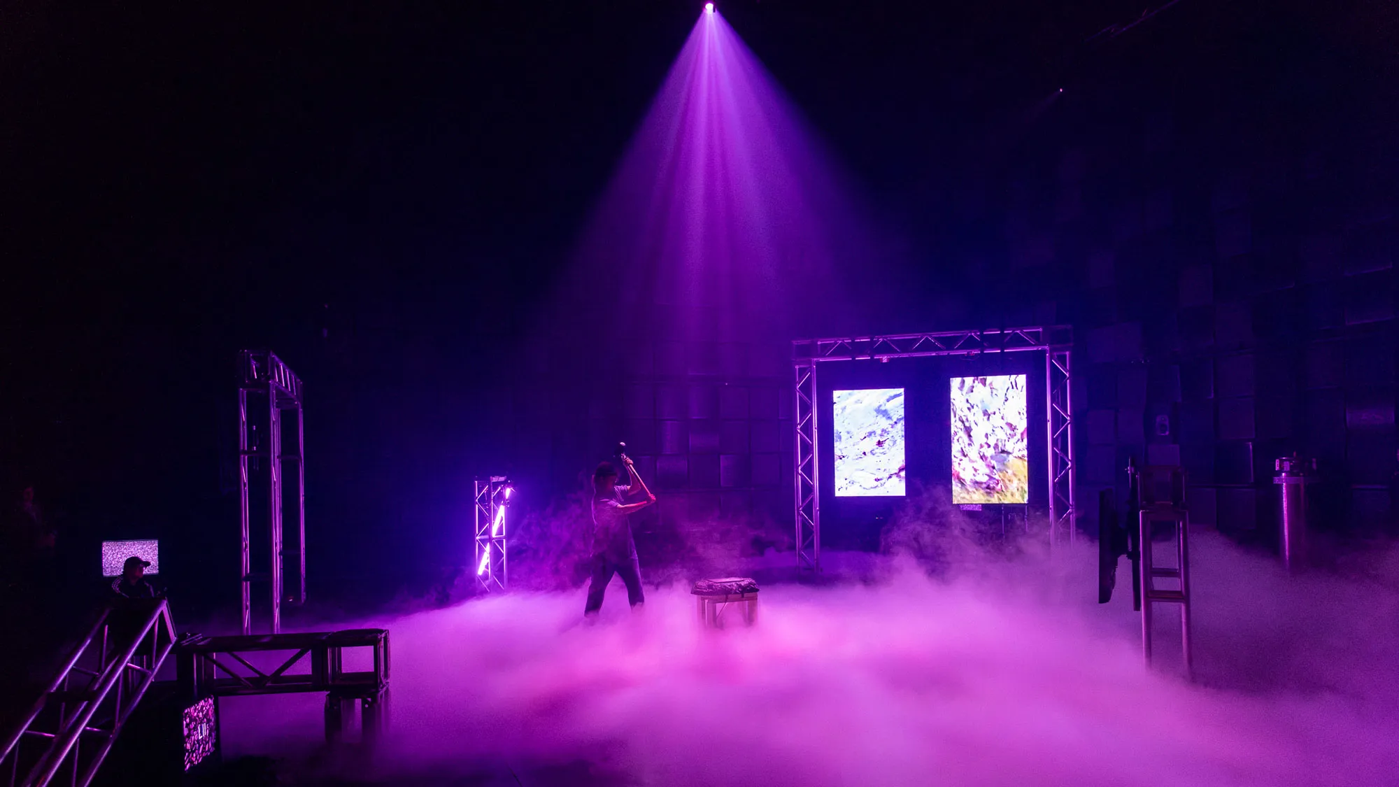 a woman strikes a surface with a five-pound hammer to tigger events in a studio lit in purple with theatrical fog rolling across the floor