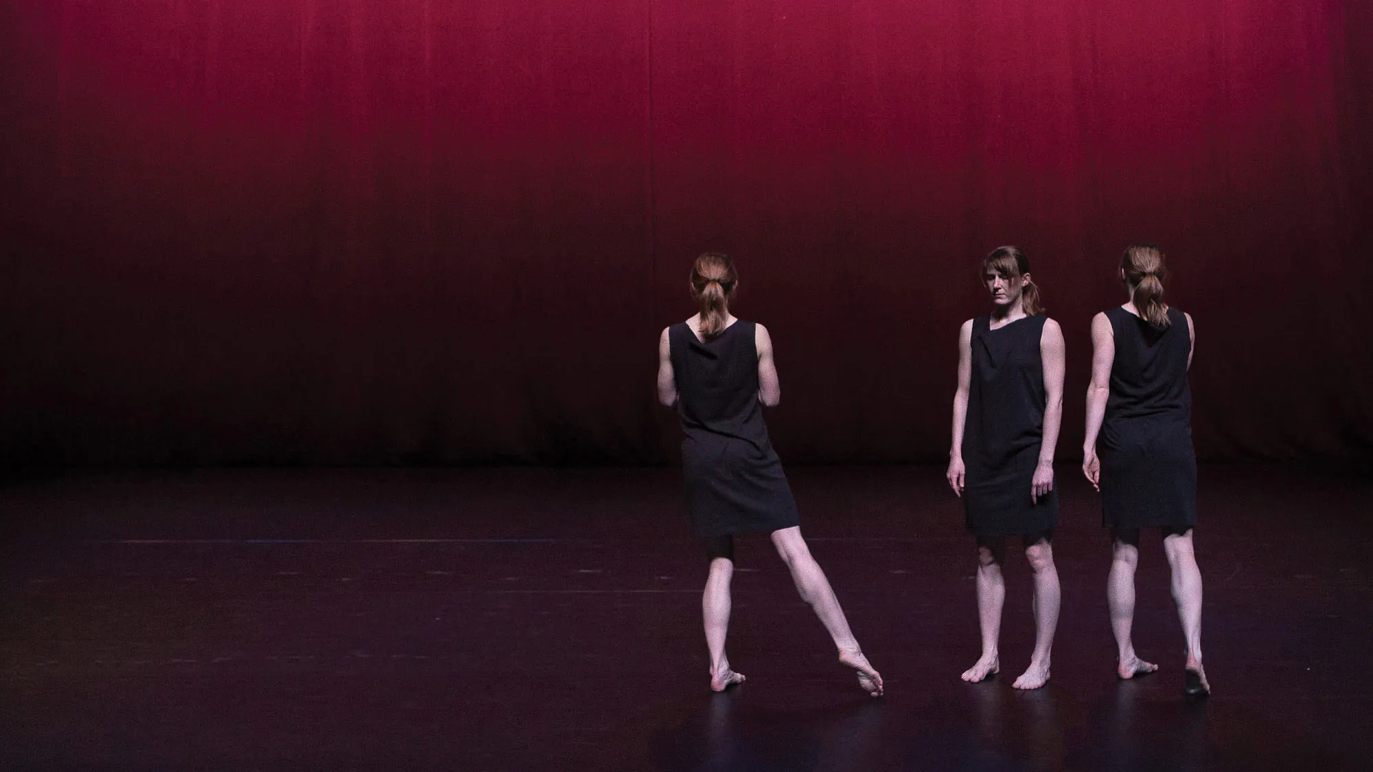 Three female dancers wearing black one shoulder dresses in a tight formation on a stage lit softly in burgundy light. 