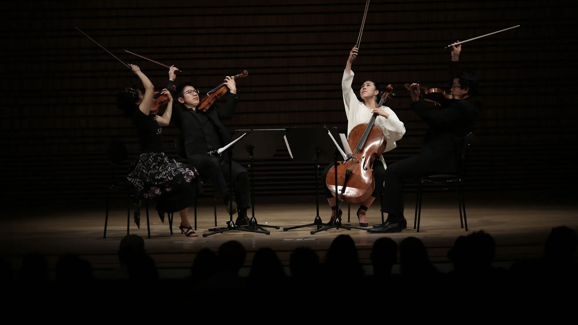 Formosa Quartet with bows lifted and pointed to the ceiling on a dimly lit concert hall stage. 