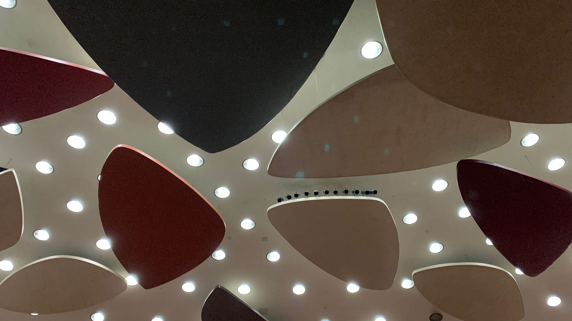 Rounded triangular geometric shapes in muted primary colors mounted on to a white ceiling with rounding lights in between. 