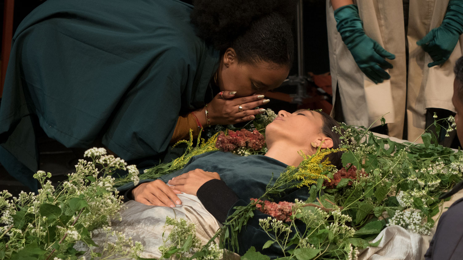 A woman laying in a coffin covered in flowers while a another woman wearing a green cloak whispers in her ear. 