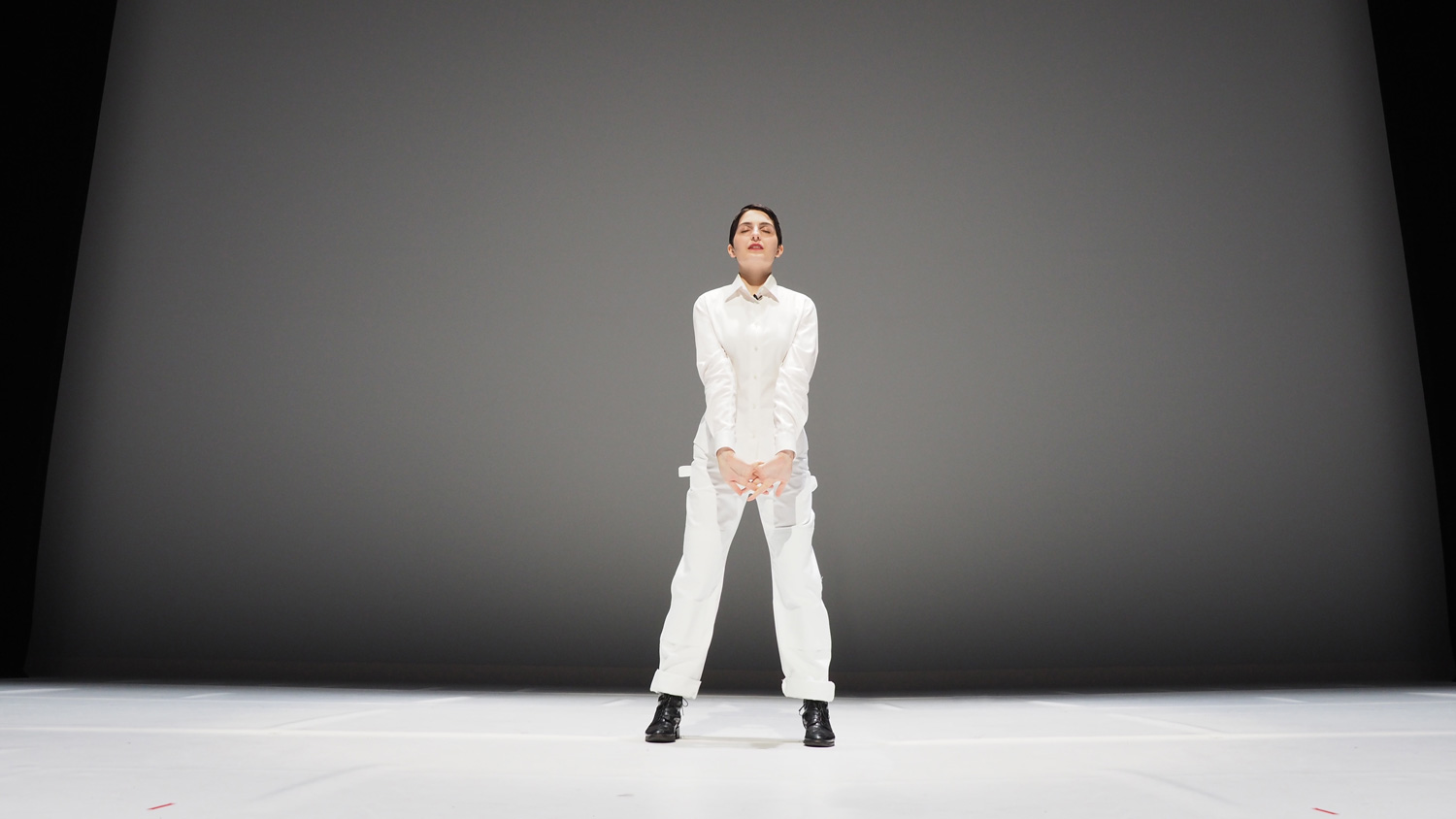 A woman wearing a white jumpsuit standing in front of a grey backdrop. 