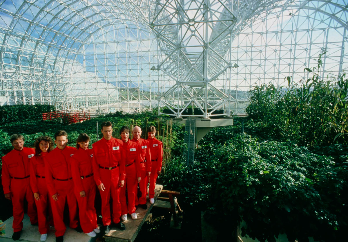 A group of people in red jumpsuits in a V formation posed inside of a biosphere filled with lush greenery. 
