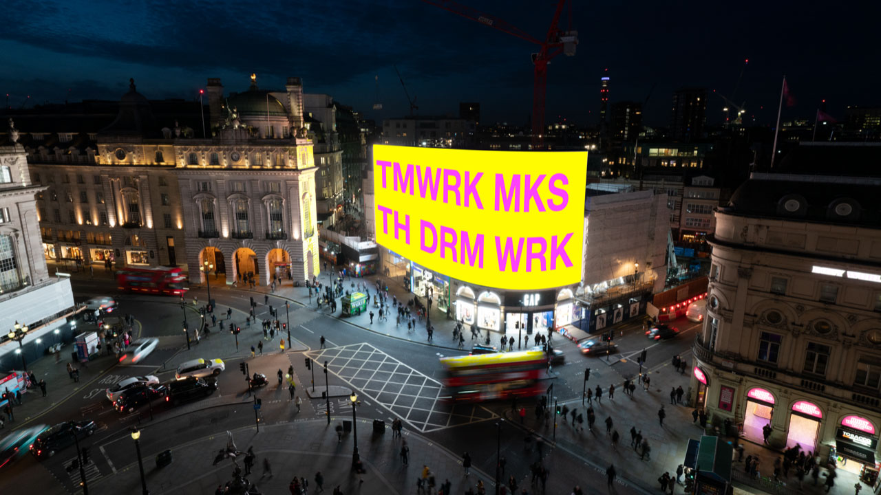 a large projection on a bright yellow screen in a town square reading TMWRK MKS, TH DRM WRK 