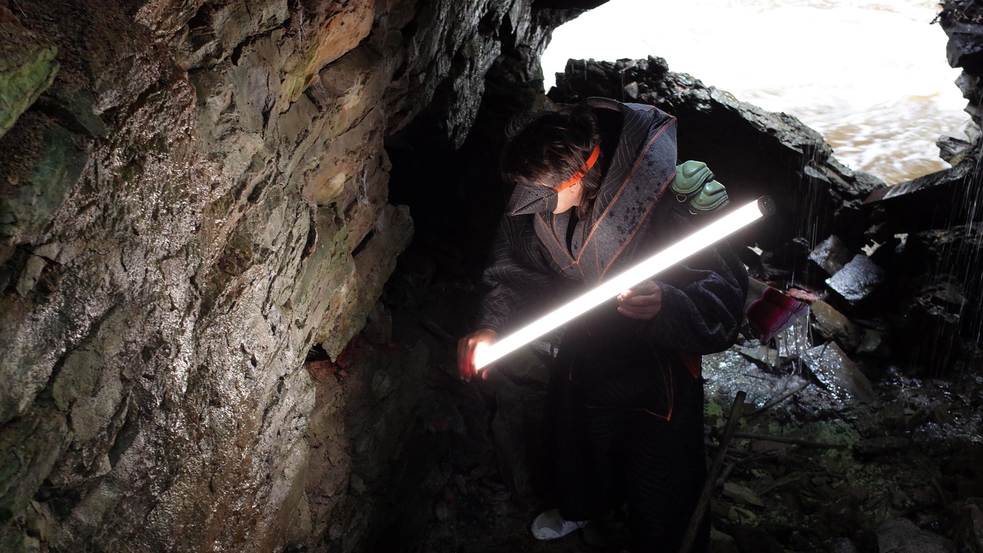 a man in a samauri-like costume with a lightsaber in a cave above a river