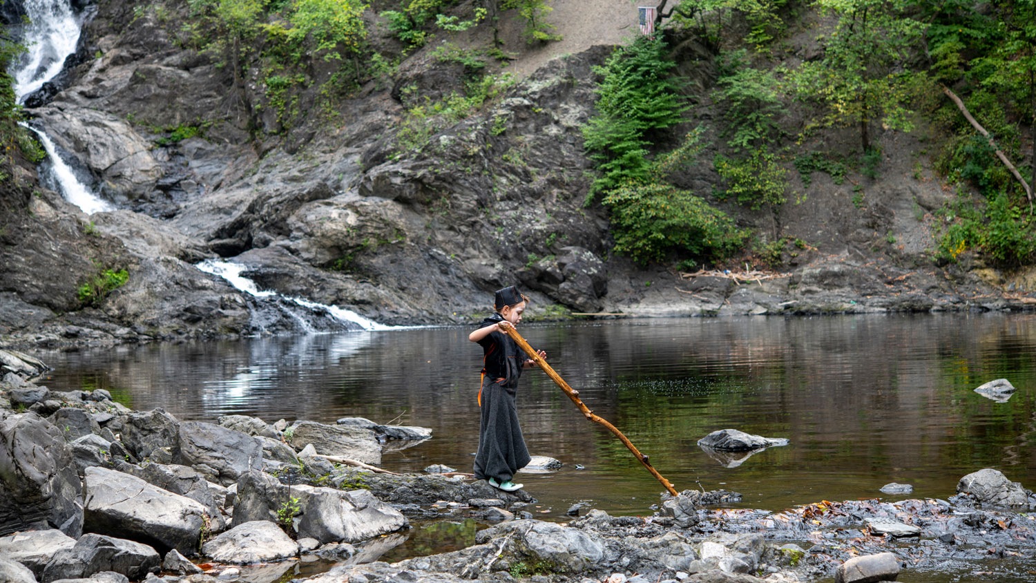 a young boy points a large stick at the rocks at the bottom of a waterfall in a deep gorge.