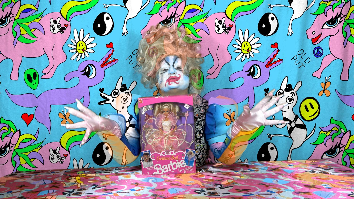 A drag queen sits at a table in front of a crazy unicorn background with a costume ball barbie between her gloved hands with long nails.