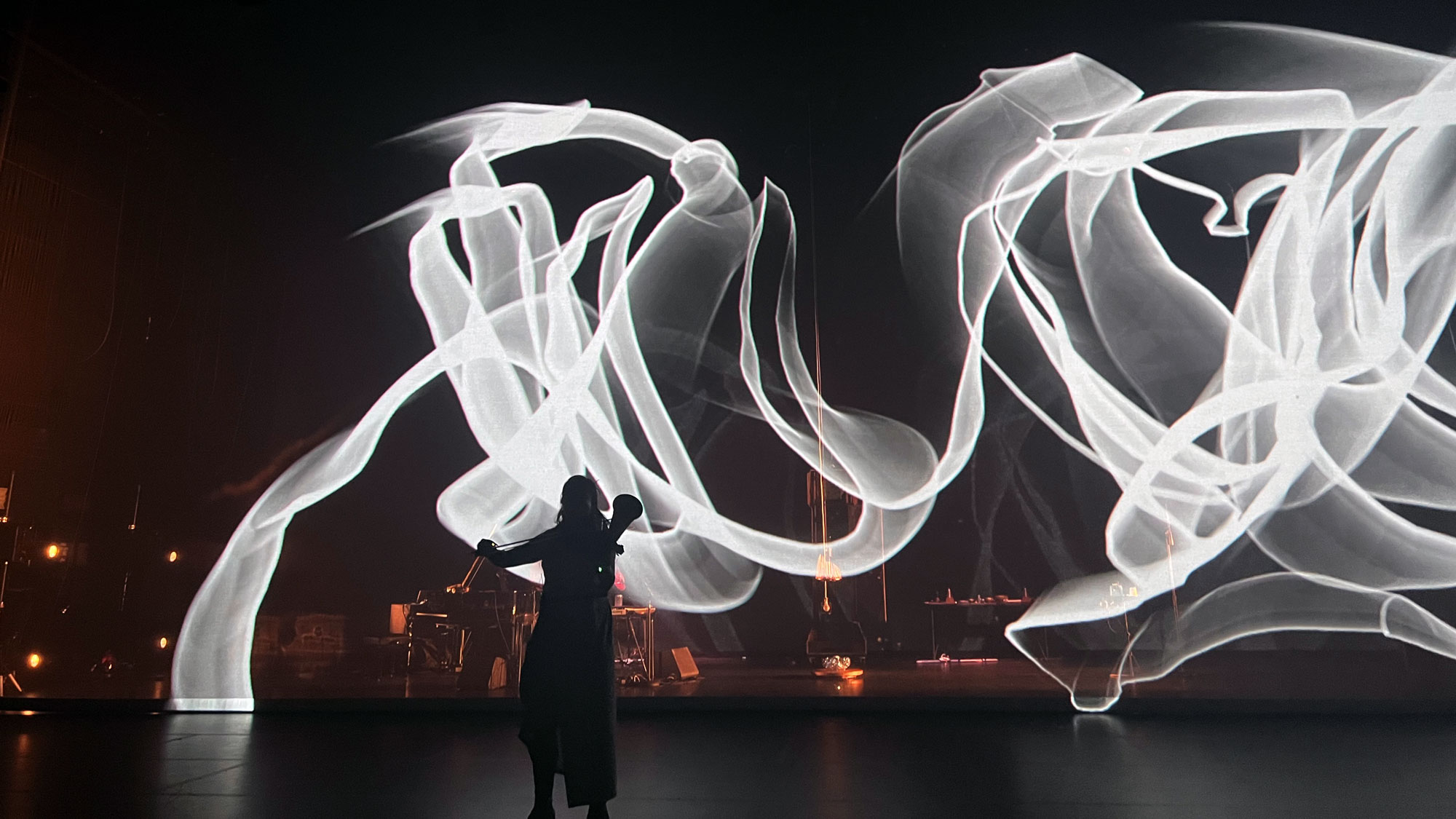 bora yoon playing their violin in the foreground with a abstract projection of ribbon-like white light in the background.