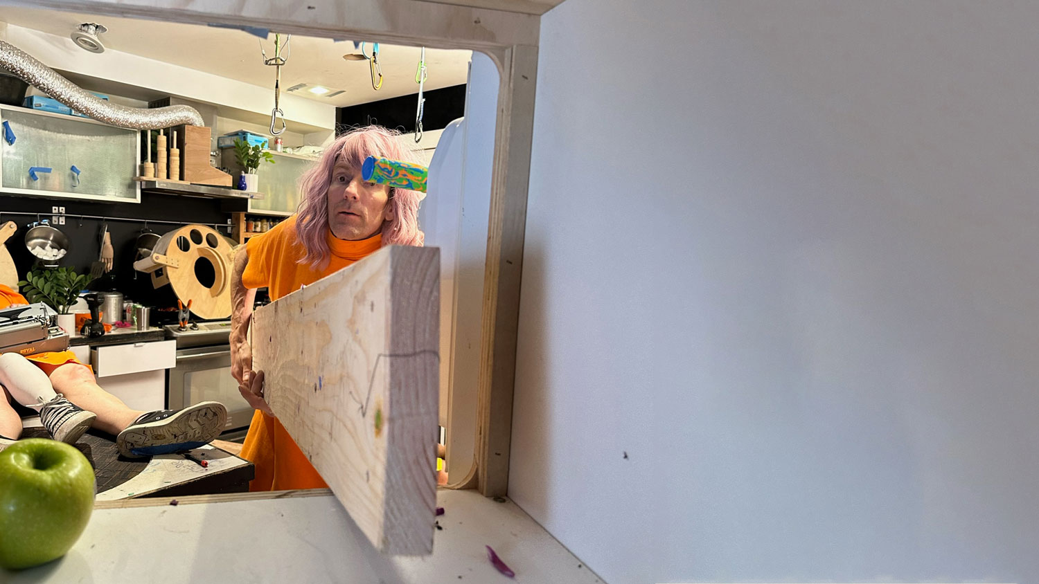 a person in an orange shirt with medium long blonde-lavender hair pushing a 2x8 pine board toward the camera. a granny smith apple sits in the left bottom corner of the frame a kitchen in the background.