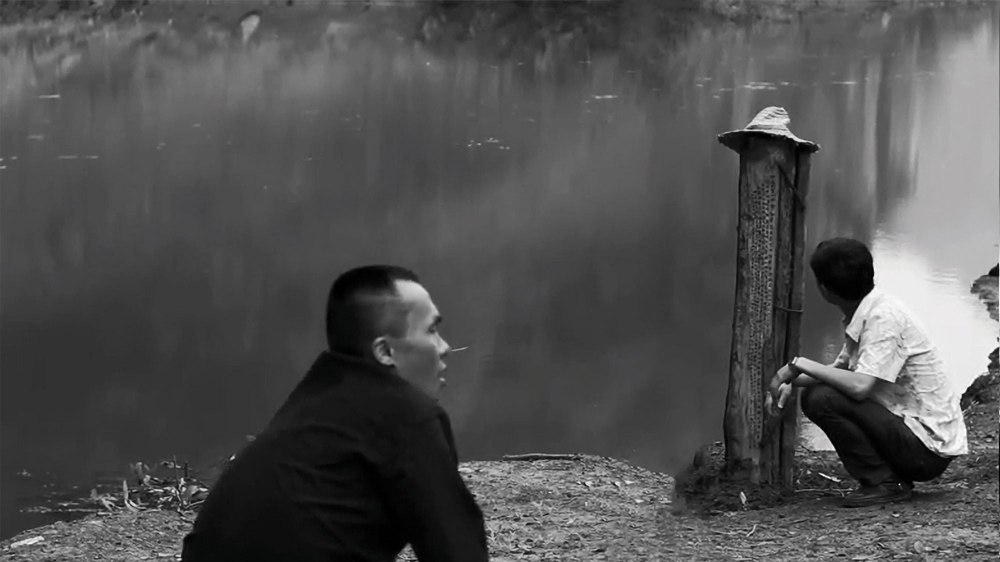 two asian men squat next to a still water body.