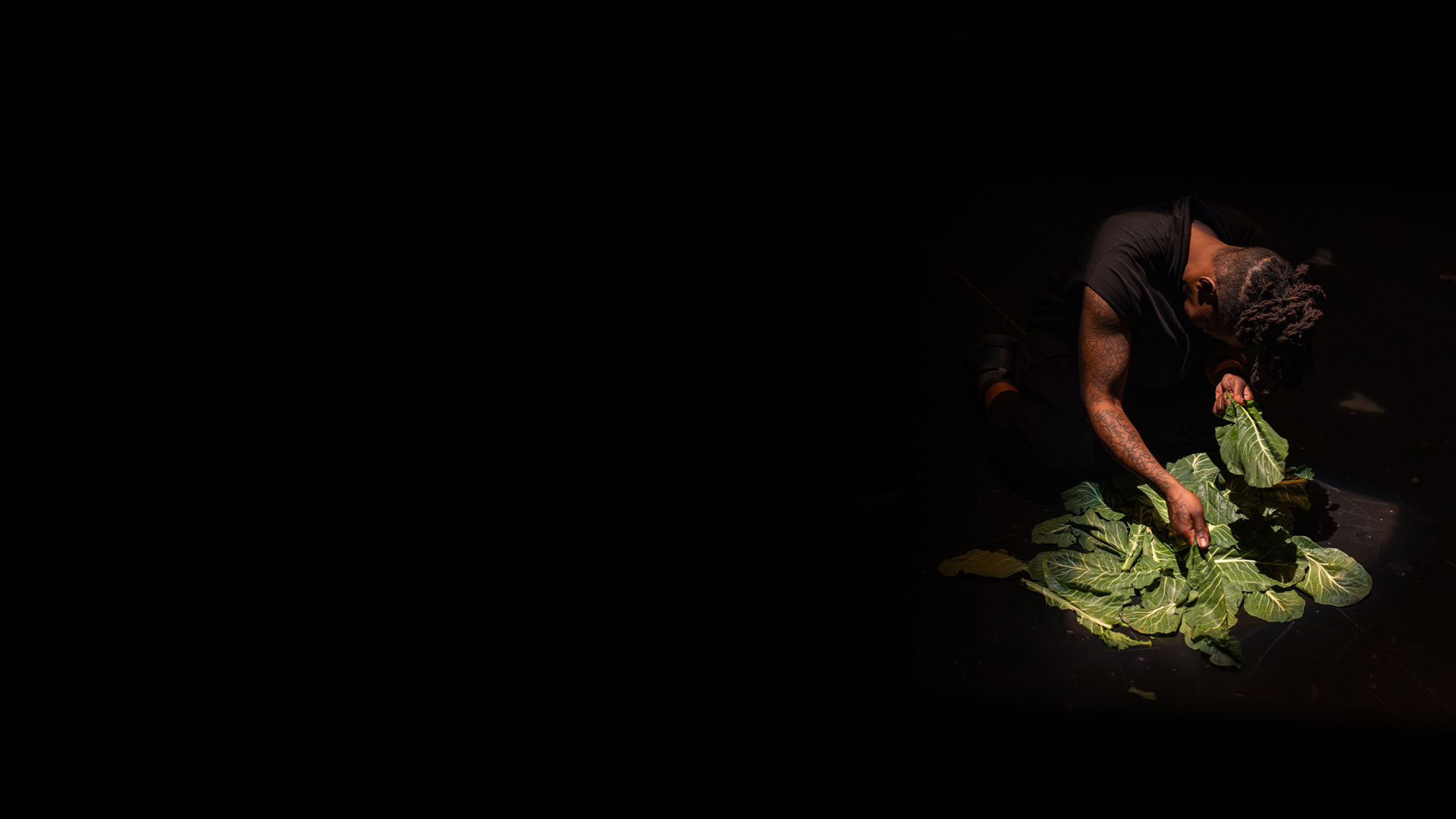 a black person keeling over a pile of collard greens in a dark venue 