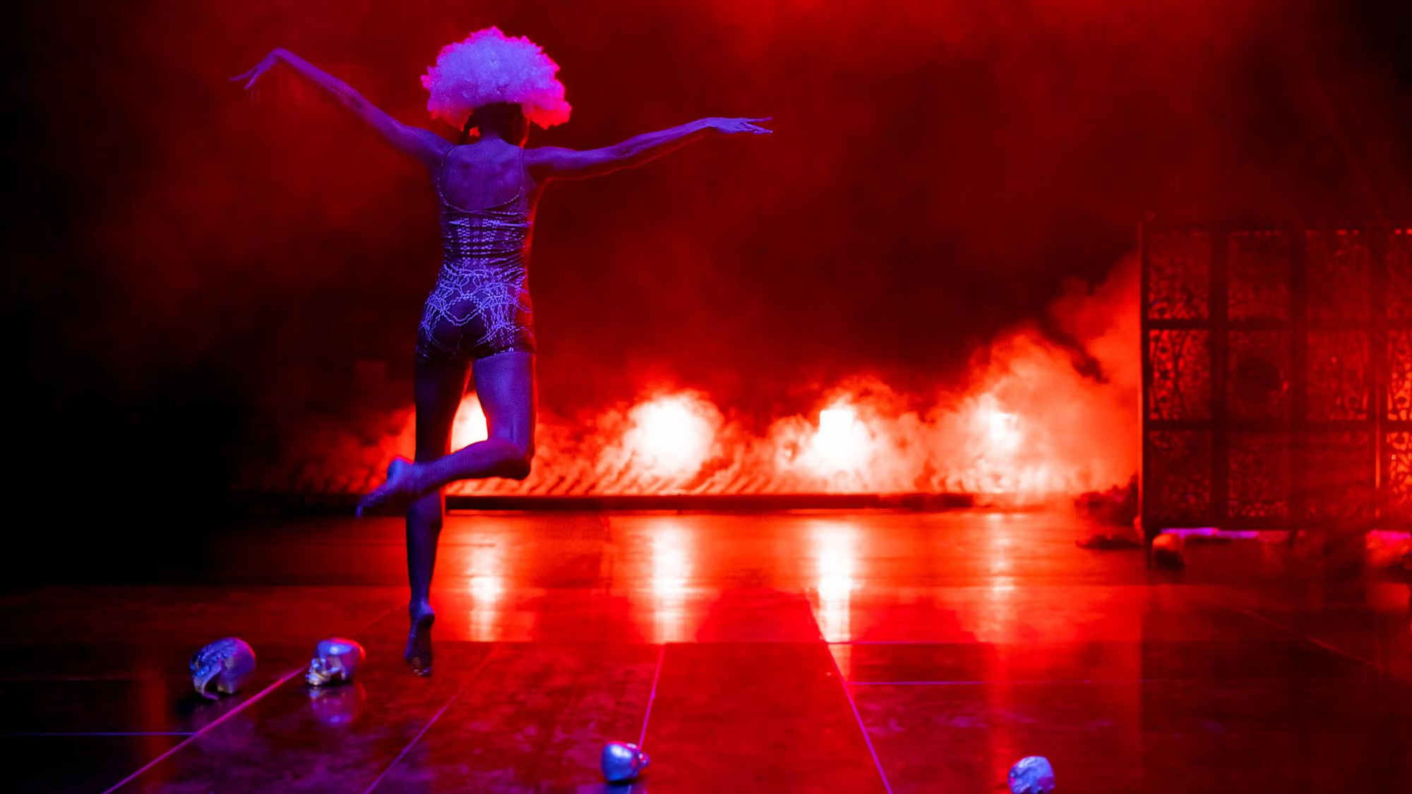 a woman runs toward the back of a stage her arms spread like an eagle, bright fire-like floor lights and fog in the background