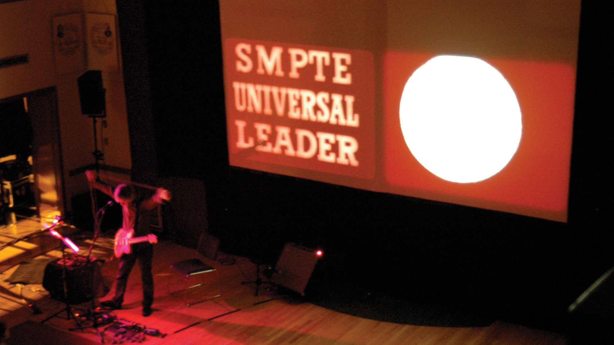 A musician standing with arms up on a small stage lit in red light in front of a projection reading "S M P T E Universal Leader" 