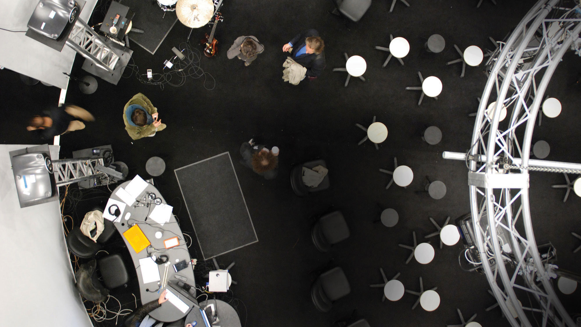 An aerial view looking down into a small, circular room with a black floor and gray walls. Gray and white round stools are set up though out the space along with two cluttered desks as folks mill about.