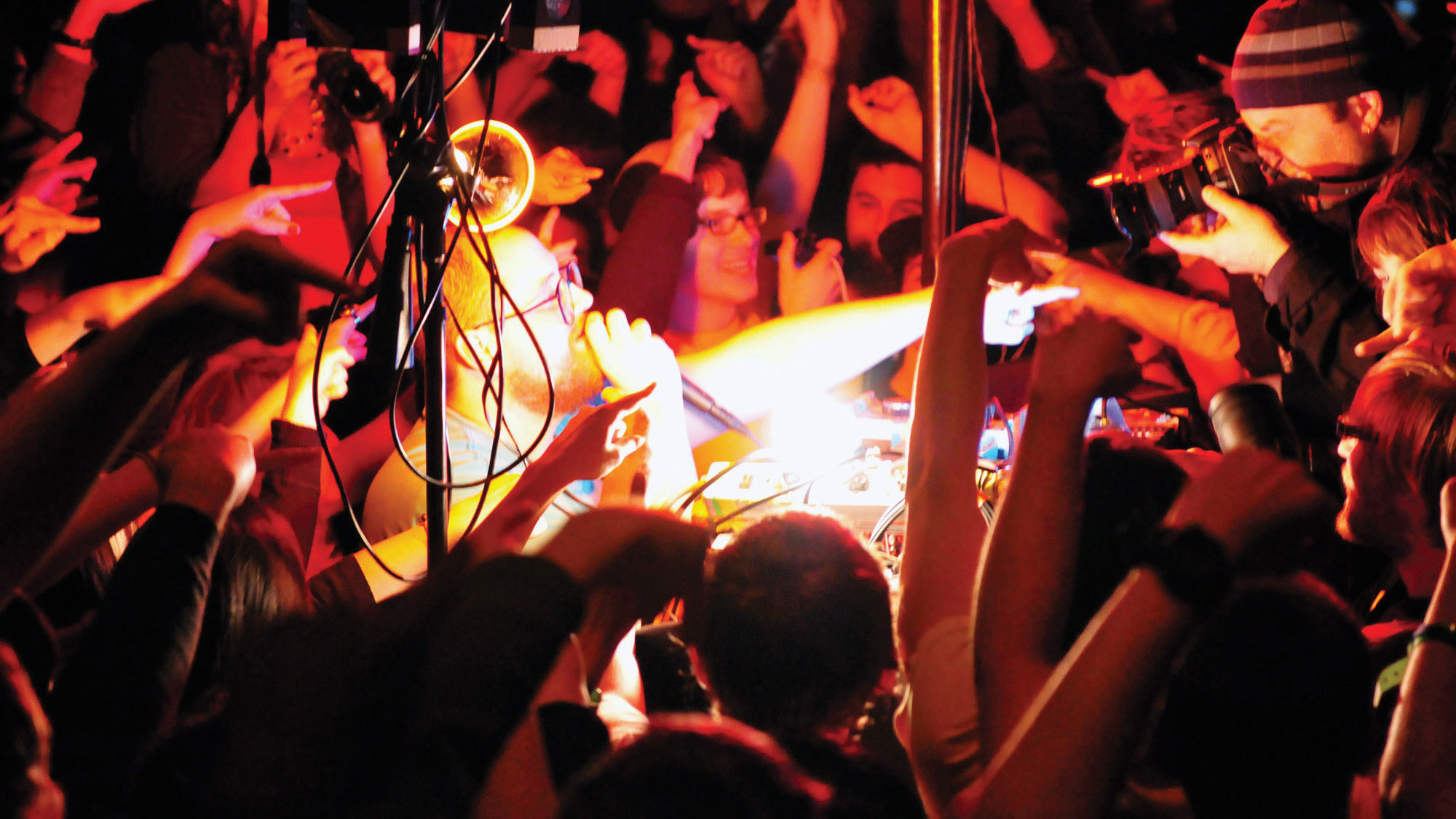 Dan Deacon performing in the middle of a tightly packed crowd, all pointing in the same direction. A beam of yellow light over exposes the middle of the scene while the rest is lit in red light. 