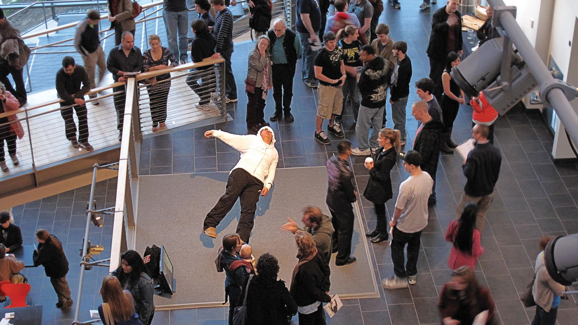 People gathered in the mezzanine watching a performer wearing a white hoodie and black pants leaning back dramatically, almost falling over. 