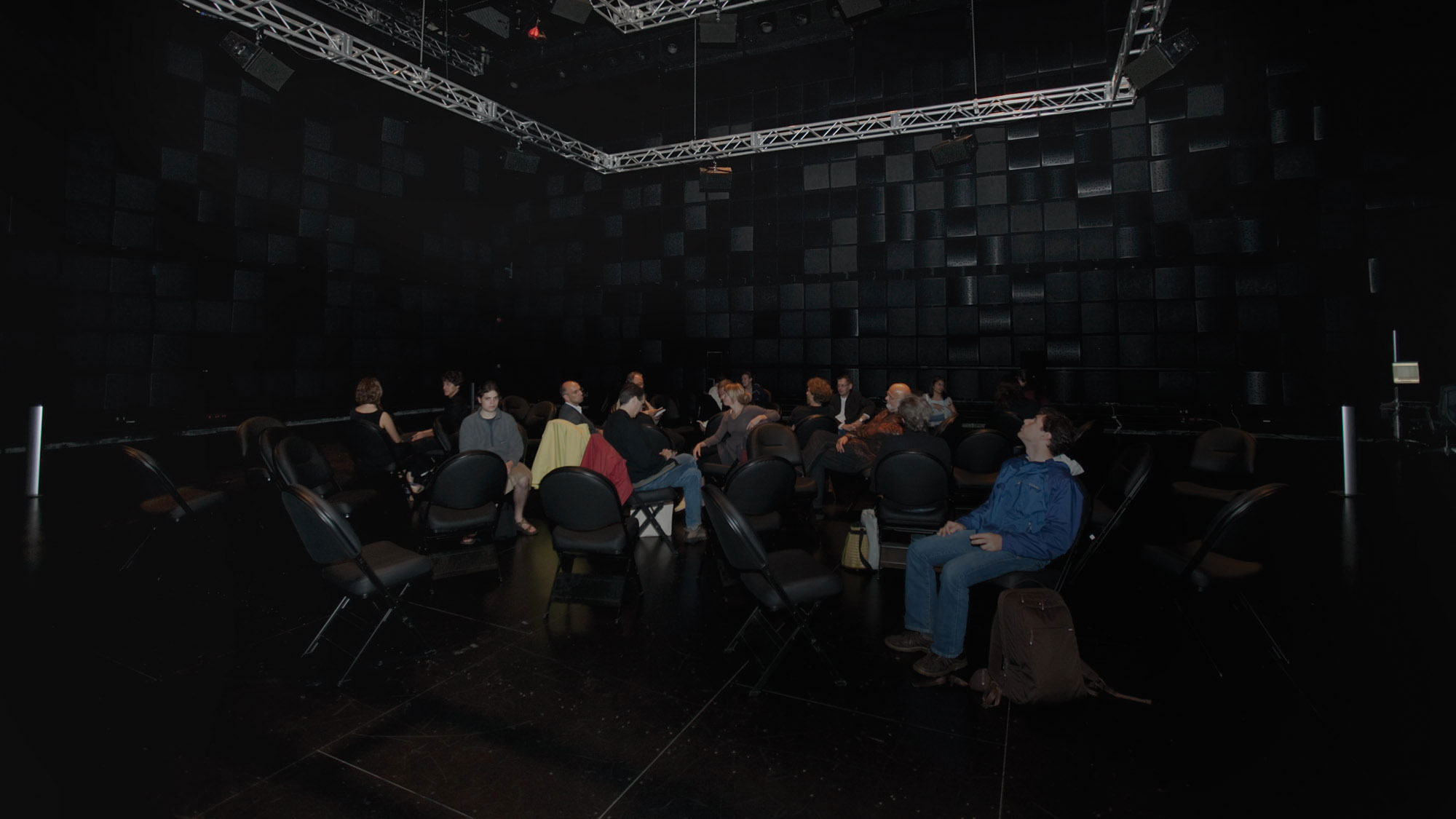 A small audience seated in black folding chairs randomly placed through a black box studio, seemingly listening to the space. A metal rigging style is suspended from the ceiling. 