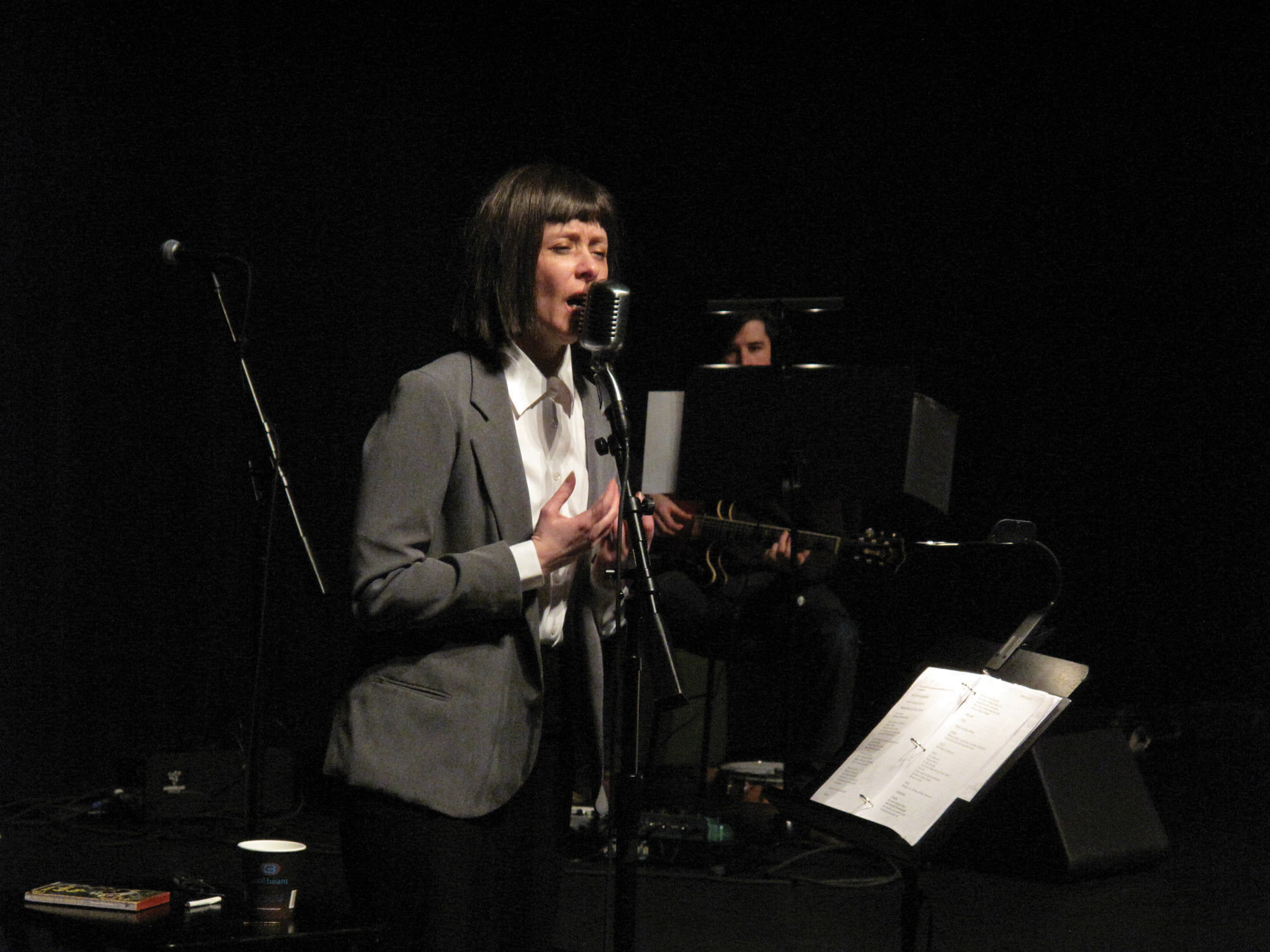 A women in a gray blazer and white collared shirt singing into a microphone. 