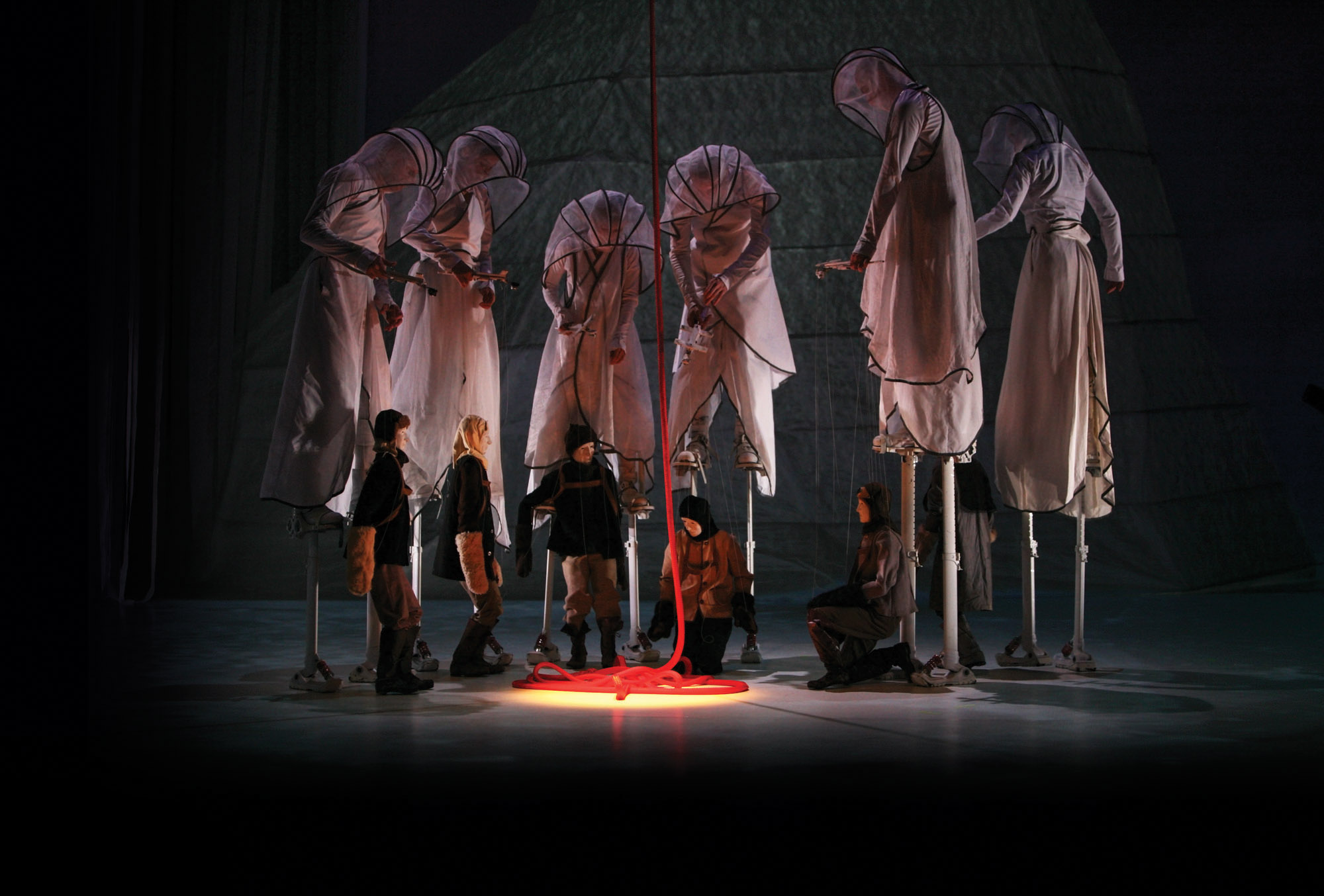A group of six puppeteers dressed in hooded white robes on stilts working large marionettes gathered around a lit red rope. 