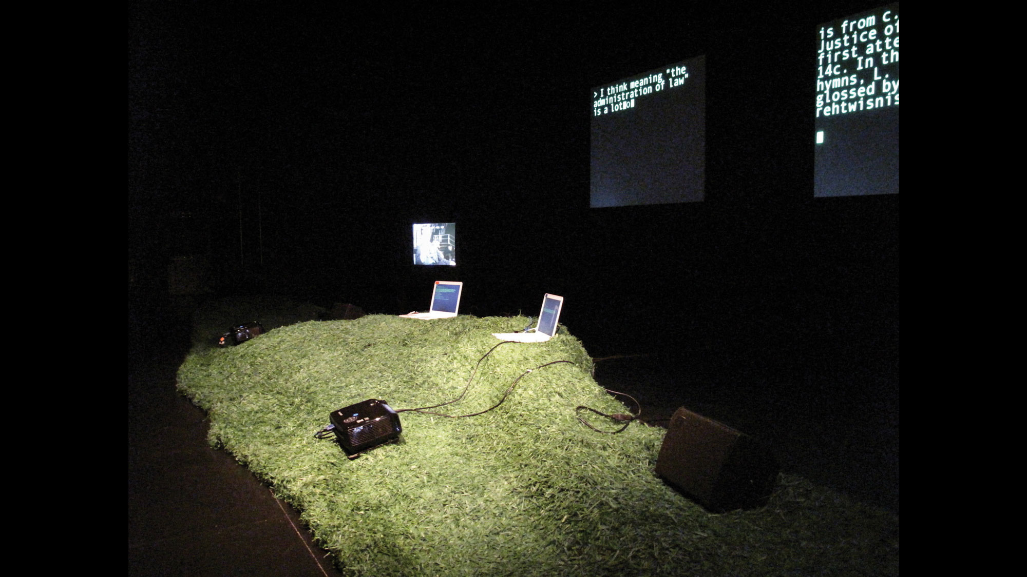 Two laptops sitting on an artificial grassy hill in front of two screens projecting blocky random text in a black box studio