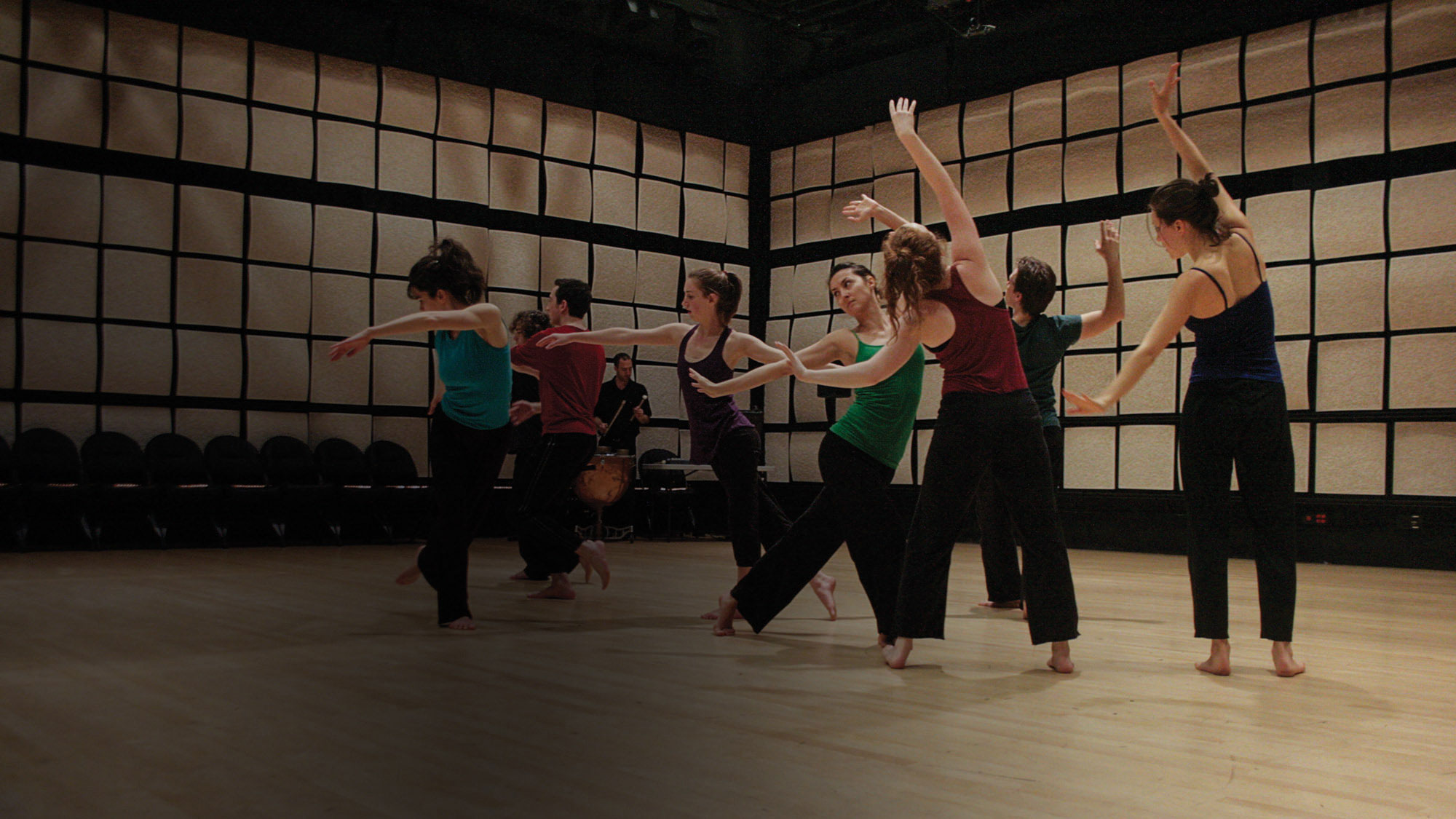 Eight dancers wearing jewel toned tops with black bottoms, moving with arms outstreched in a room with tan acoustic tiles on the walls. 