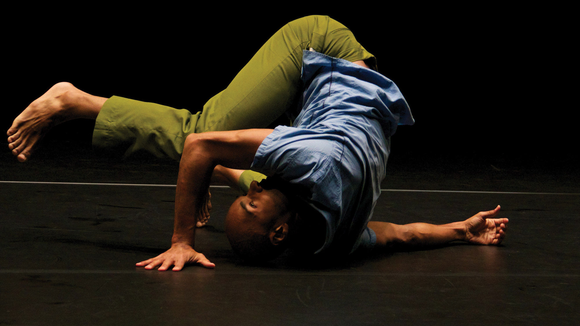 A Black man wearing a blue top and green pants laying on the stage floor with his leg over his head. 