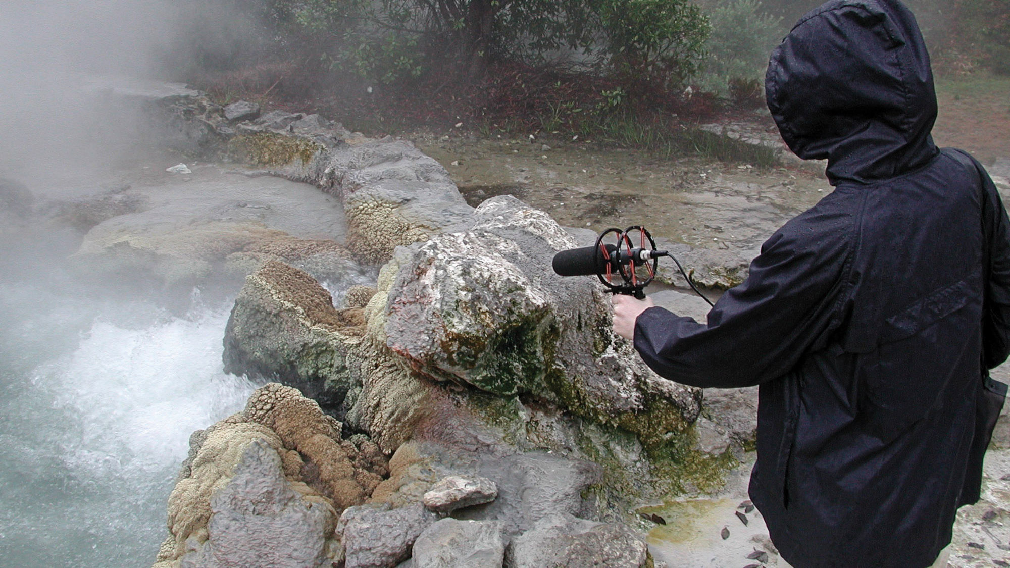 A person in a navy blue rain jacker with hood up holding a microphone over some natural hot springs. 
