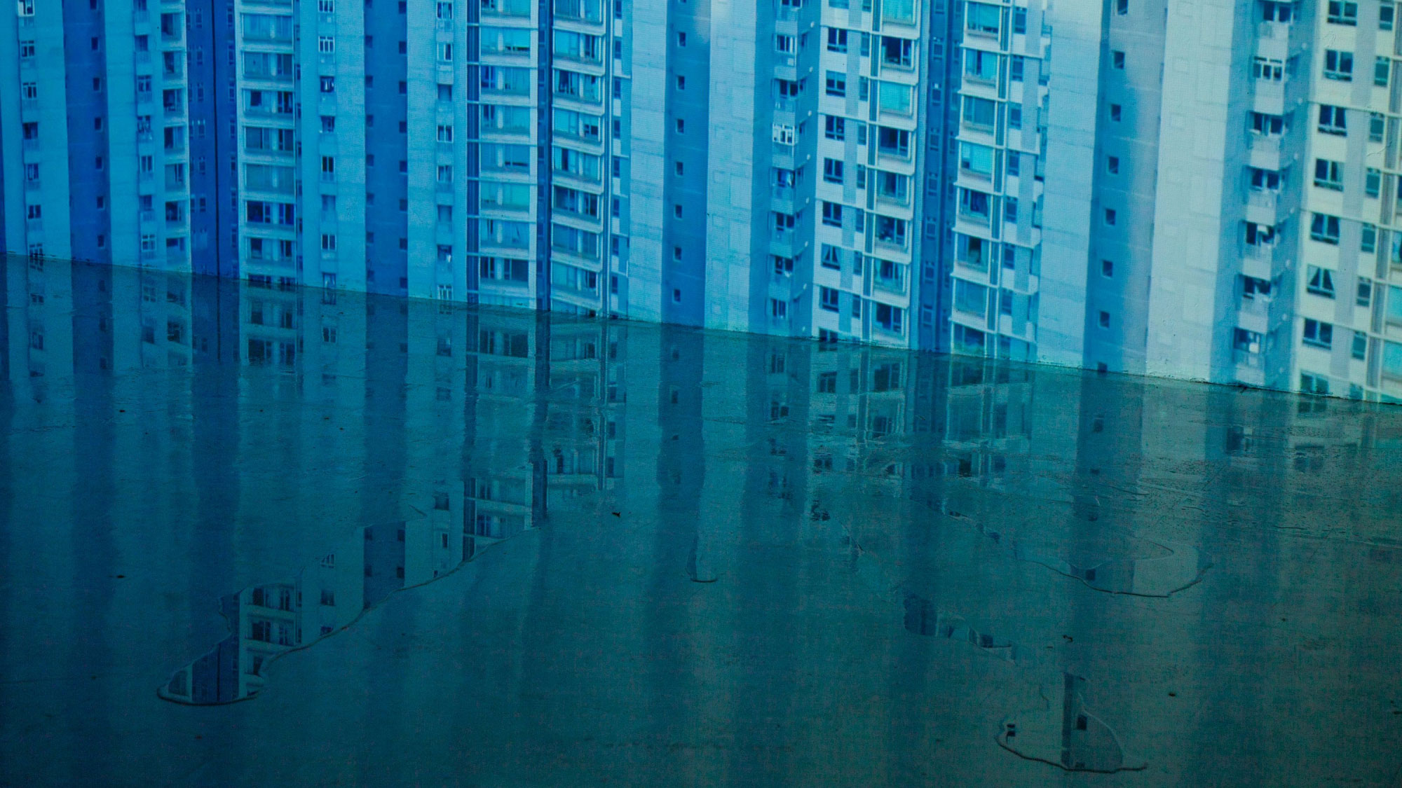 A blue high-rise apartment building with mean windows projecting on to a wall. A small puddle forming a reflection is on the floor where the wall meets the ground. 