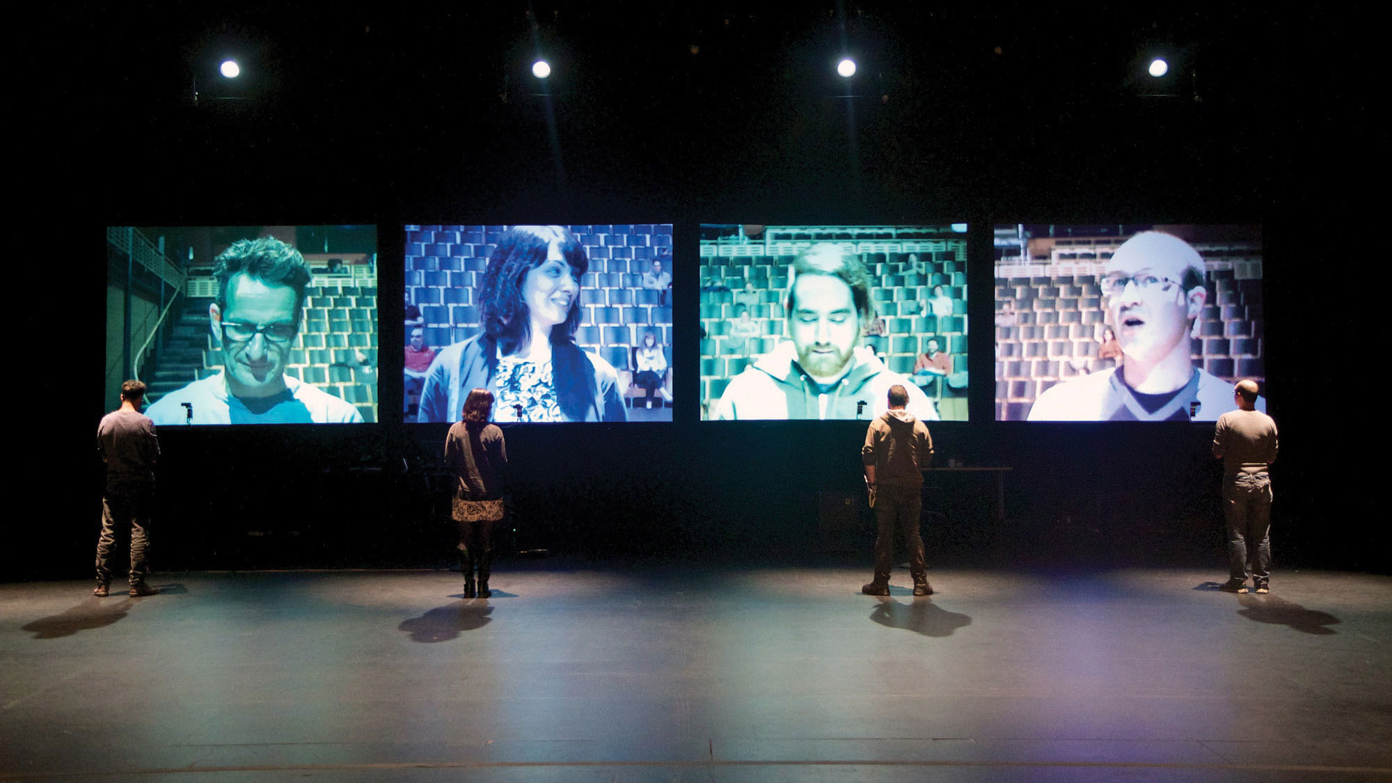 Four people standing on stage, back to the viewer looking at a projected image of themselves on a four screens in front of them. 