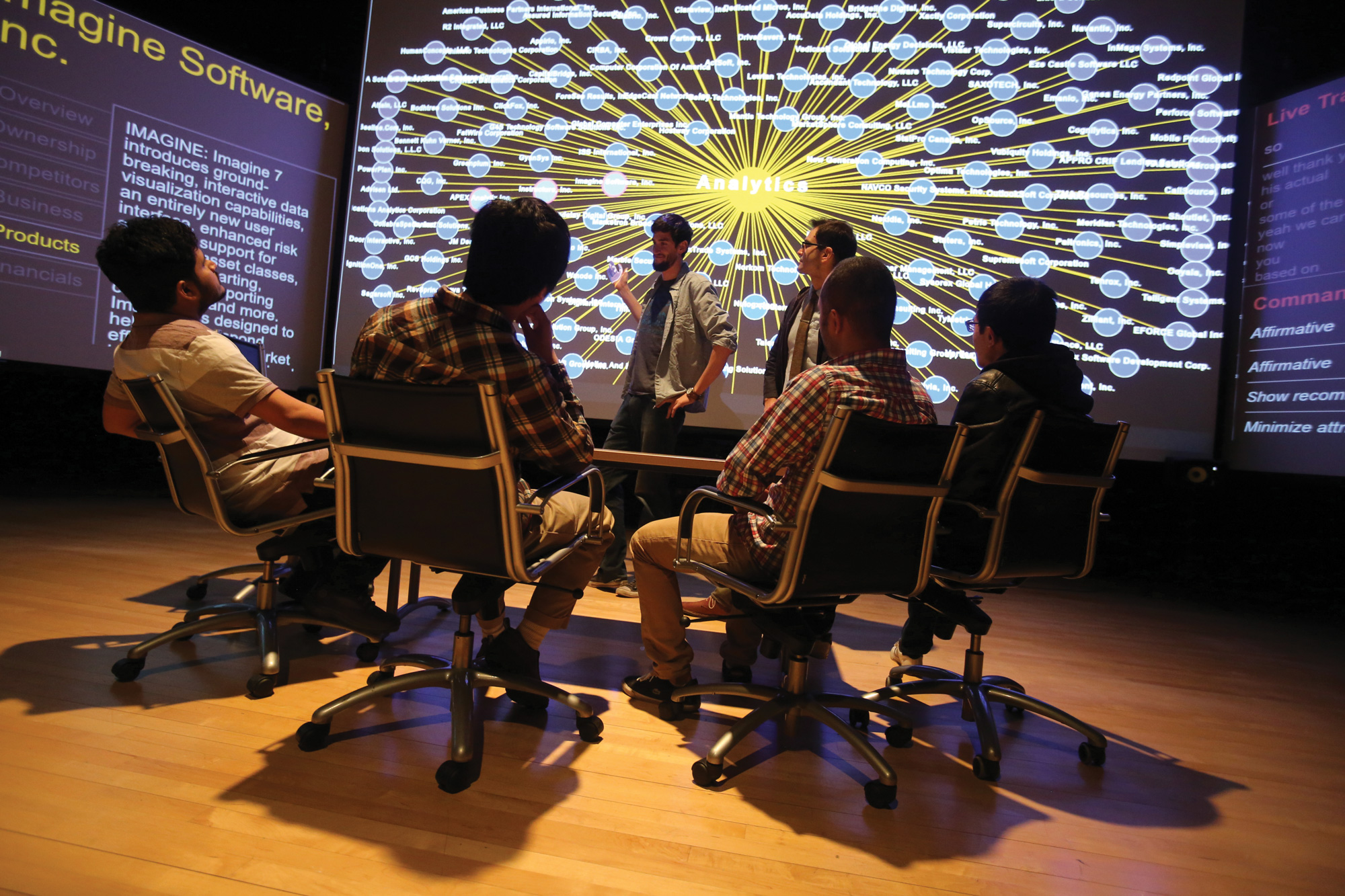 a group of four students facing two students giving a presentation in front of large screens with projections on them