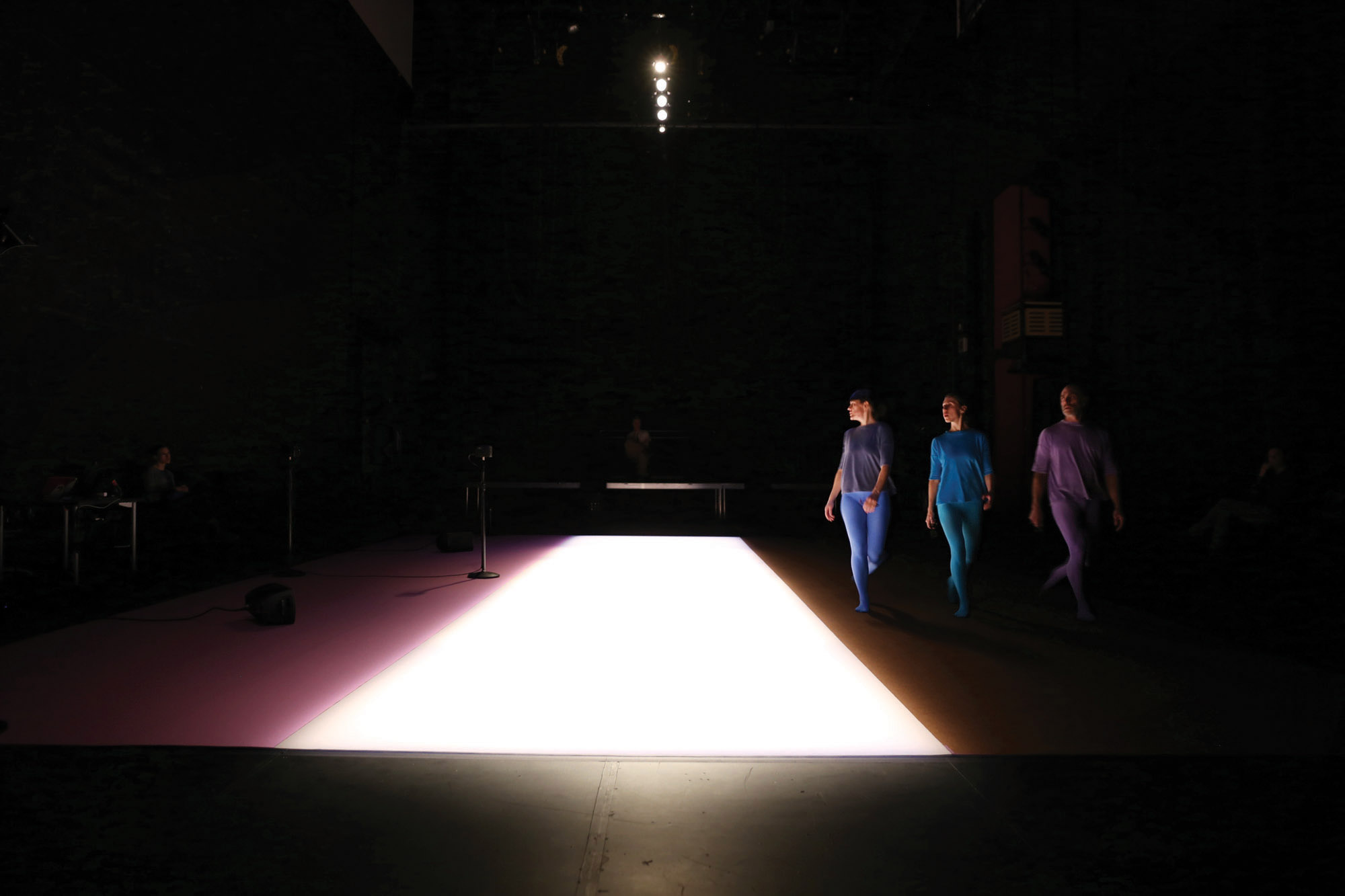 Three dancers wearing periwinkle, blue, purple walking around a rectangle of light on the floor