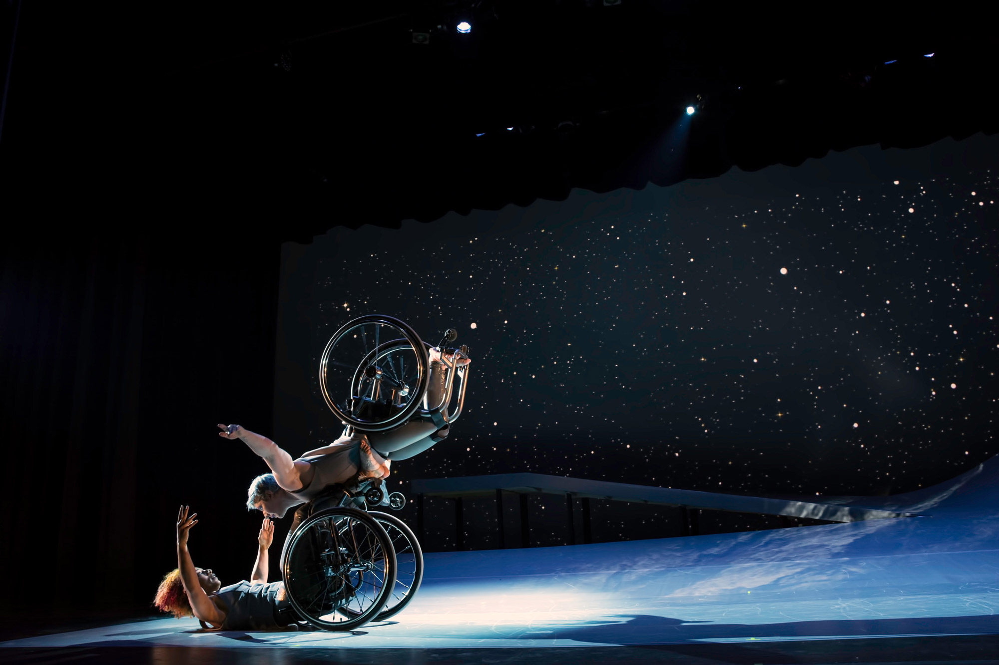 A white woman with short hair in a wheelchair flying in the air with arms spread wide, wheels spinning, and supported by a black woman also in a wheelchair with curly hair who is lifting from the ground below. 