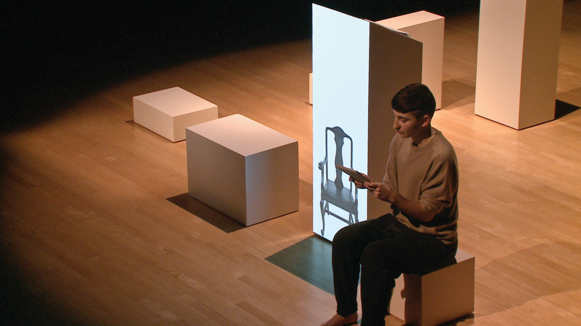 Gordon Hall sitting on white cube next to a projection of a chair on to a larger white cube 