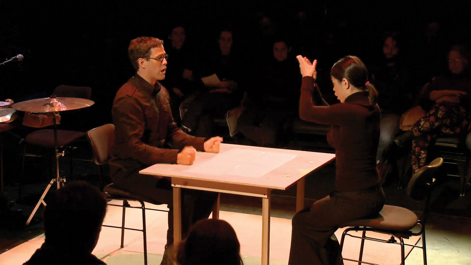Two people seated at a small square table on a stage. The woman has her hand in a clapping motion above her head. The man looks t hr with mouth open and hands on table in fists. 