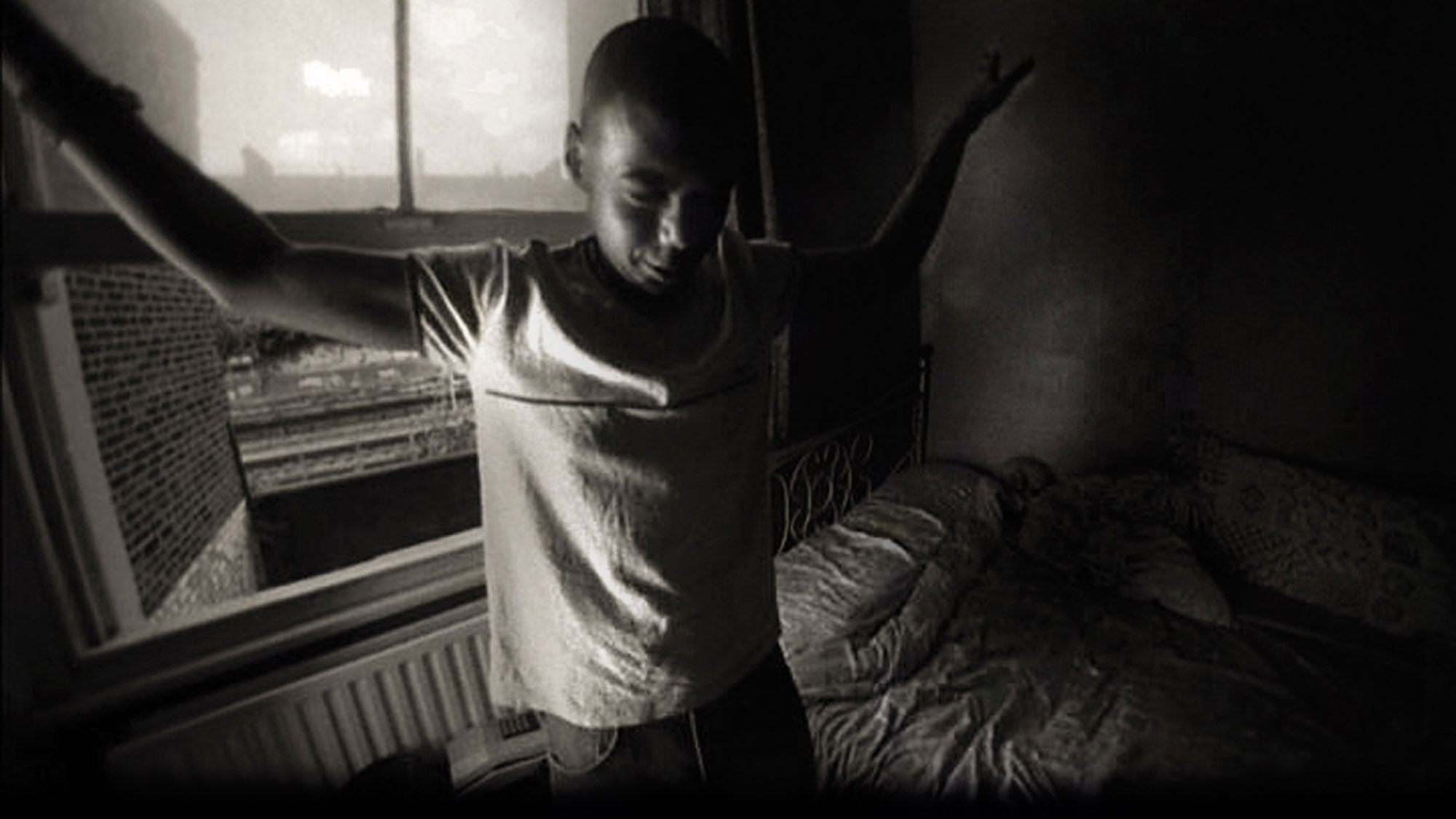 A young Black man standing in an apartment bedroom next to an unmade bed with arms outstretched to the sides. 