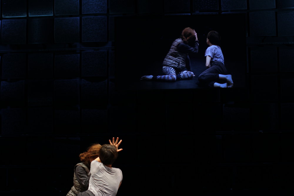 Two people in a black box studio reaching up towards a projection of themselves. 
