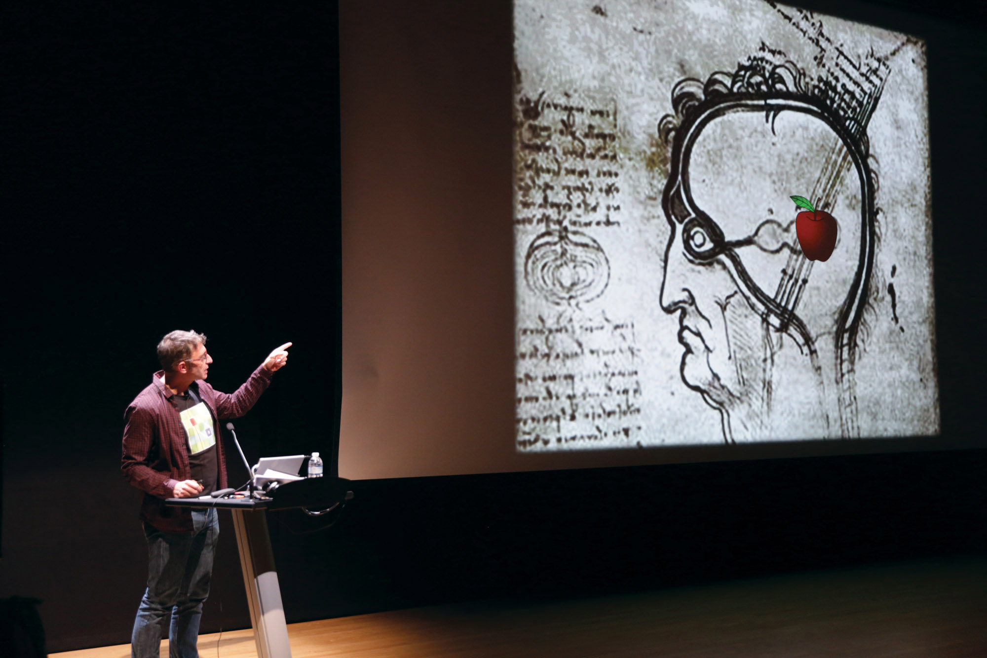 A man on stage pointing at a large screen to his left that is projecting a profile of a man, drawn by Leonardo da Vinci with an emoji apple 