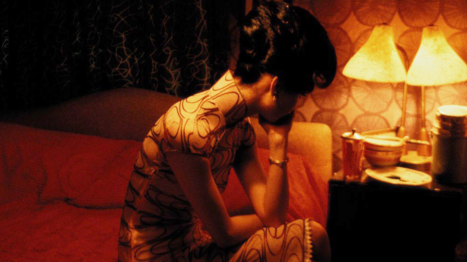 An asian woman wearing a Cheongsam sitting on a bed in an orange lit room with her head in her hand looking forlorn. 