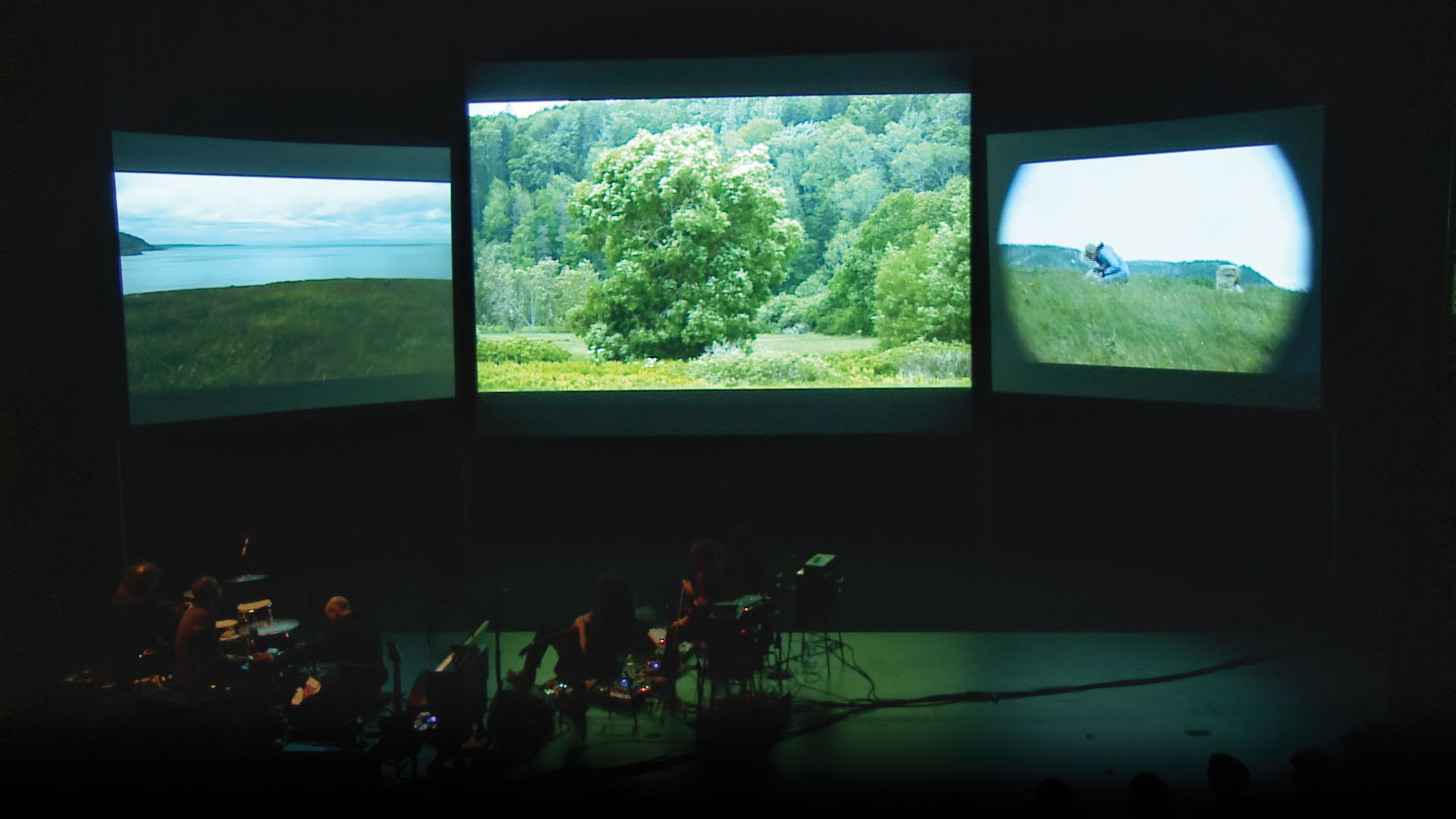 Three screens showing greenery and rolling hills behind a small pit orchestra on a dark stage. 