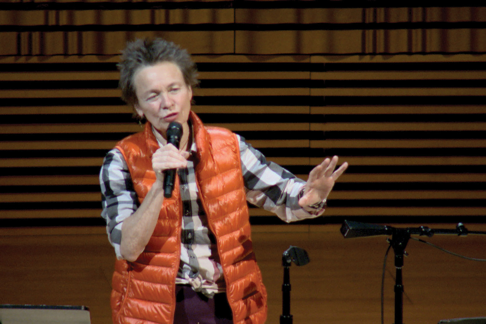 Laurie Anderson wearing a black and white plaid shirt and orange vest speaking into a microphone. 